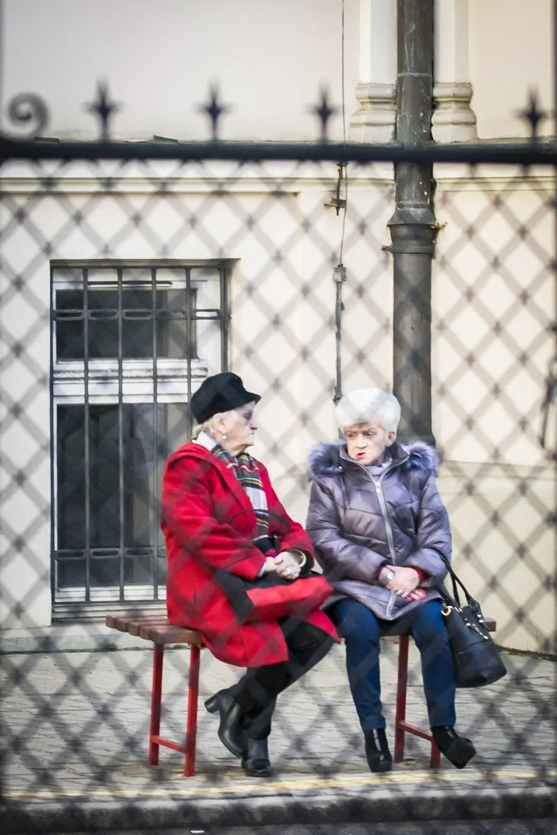 Two elderly women in colorful coats sitting by an iron fence